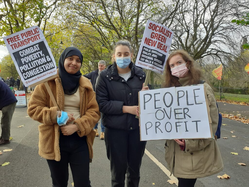 Three young women activists with SSP placards reading "free public transport", "socialist green new deal", and "people over profit"