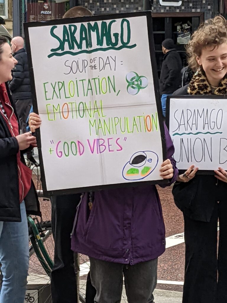 A young protester holds a sign reading "Saramago soup of the day: exploitation, emotional manipulation, 'good vibes'"